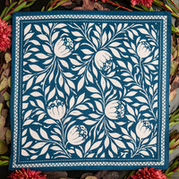 A square textile or scarf laid flat on a dark surface, featuring a white floral and leaf pattern on a deep blue ground, surrounded by red flowers and green leaves.